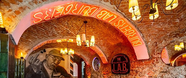 Rustic brick bar with arched ceilings, warm lighting, neon signs reading 'BURGERS', and wall art including a man’s portrait.