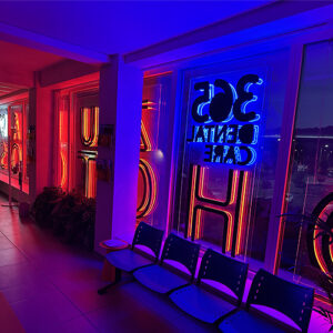 Neon-lit hallway outside a cafe or bar, with blue and red lights, window signs reflected in the glass, and a row of plastic chairs along the wall.