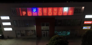 Night view of a building with red neon sign 'POHOTOVOST' above a central entrance; blue '365 DENTAL CARE' sign and other lit signs on the facade at night.