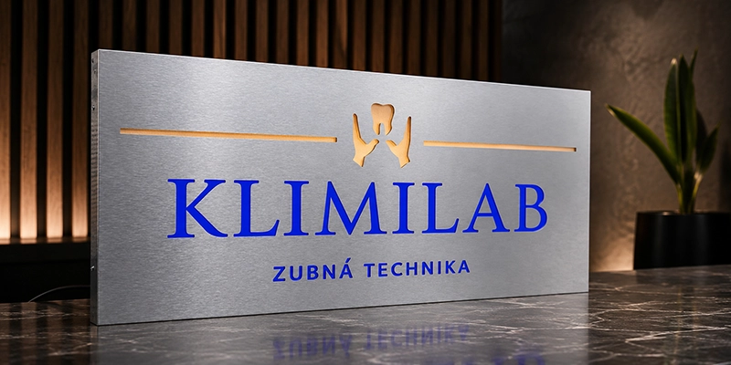 Brushed metal sign reading 'KLIMILAB' in blue with 'ZUBNÁ TECHNIKA' below and a tooth-like emblem above, on a dark reception desk.