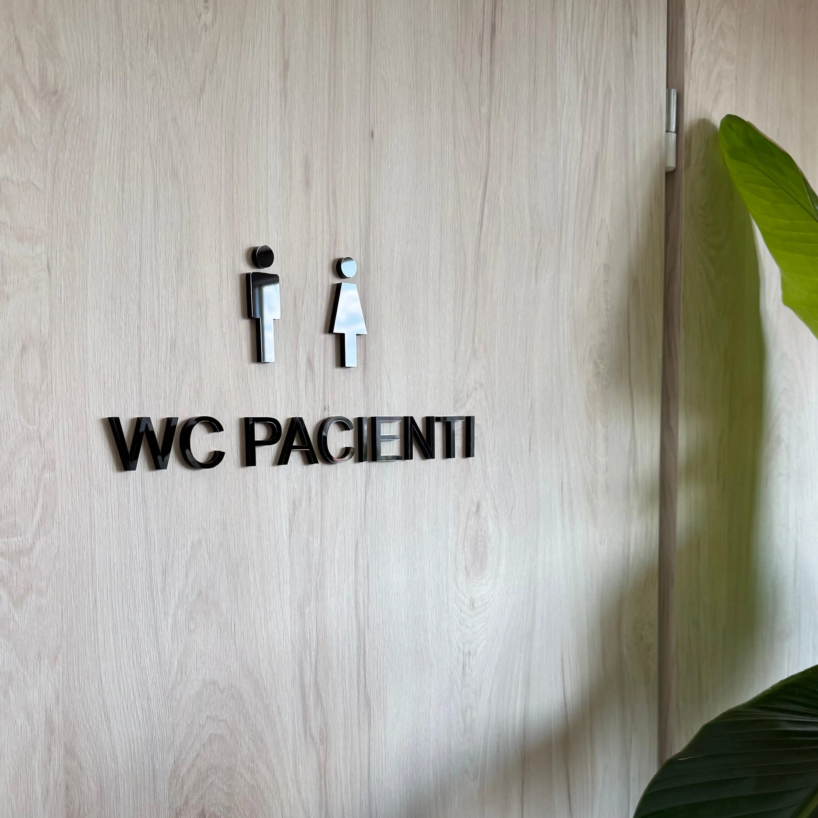 Restroom sign reading 'WC PACIENTI' with male and female icons mounted on a light wood wall, plant is visible on the right side.