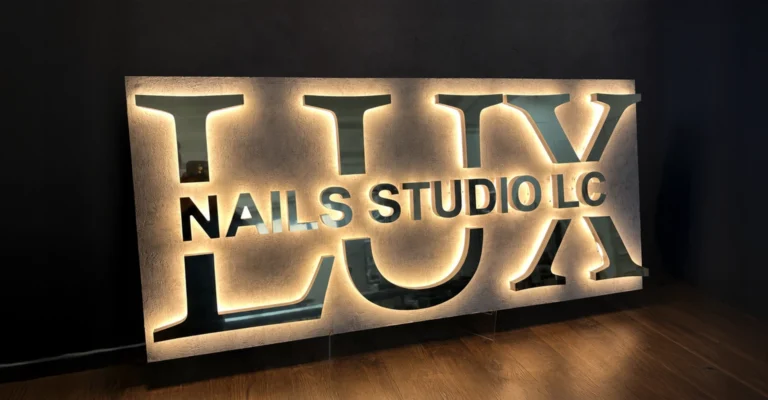 Backlit sign reading 'LUX NAILS STUDIO LLC' mounted on a dark wall, sitting on a wooden floor at an angle.