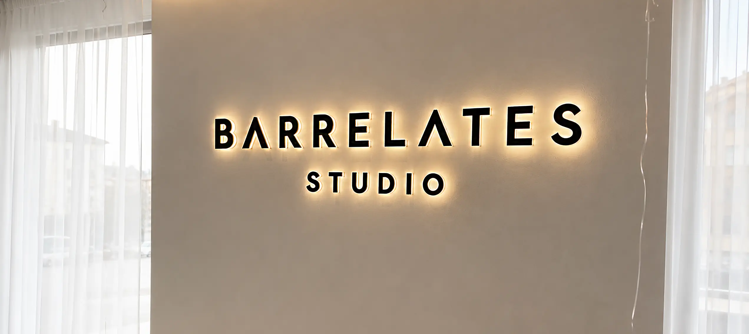 Backlit wall sign displaying BARRELATES STUDIO on a beige wall with sheer curtains nearby.